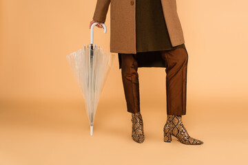 partial view of woman in leather boots and brown pants leaning on umbrella on beige background