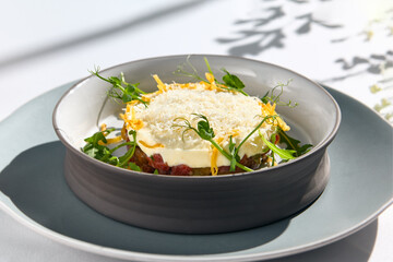 Beef tartare under cheese sauce with deep fried onion. Fancy dinning with beef tartare on white table with simple contemporary decoration. Sunlight and harsh shadow still life. Beef tartare blue dish.