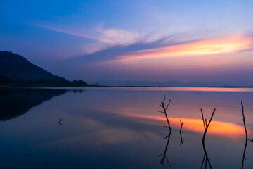 Twilight sun shines upon the clouds as a reflection of the water surface, with branches in the background, with mountains and the night sky in the background.