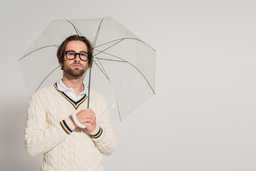 man in white knitted pullover standing under transparent umbrella isolated on grey