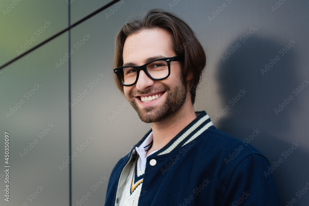 Wall mural happy young man in stylish eyeglasses standing near grey wall while smiling at camera