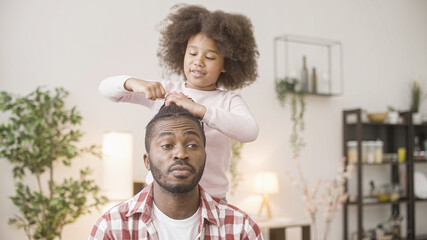 Little black girl making hairstyle for patient father, having fun, family time