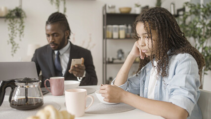Sad black teenage girl sitting next to busy father working in the morning, loneliness
