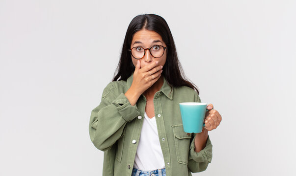 young hispanic woman covering mouth with hands with a shocked and holding a coffee mug