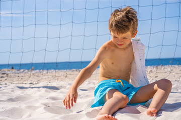 Handsome young boy in blue shorts and sunglasses sits on the white sand on the beach. Plays with white sand.