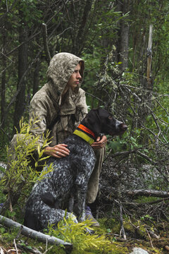 A girl sits in the woods with her hunting dog. A girl in a comuffle hunts a duck with her German drathaar