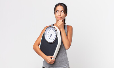young hispanic woman thinking, feeling doubtful and confused and holding a weight scale. diet...