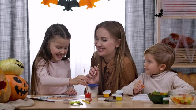 Happy caucasian woman and her two children, daughter and son, are sitting at the table, talking and drawing pumpkins together. Decorating the house for Halloween. Slow motion. Close up.