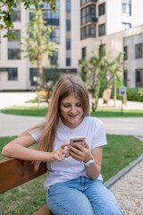 A teenage girl sits with a smartphone on the street on a summer day, smiling. She's happy. She has a video call with friends. The concept of virtual communication.