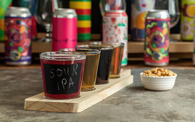 Craft beer tasting, sour fruit IPA, lager, stout, and pale ale. Bar with different cans in the blurred background. Wooden board with flight set. 
