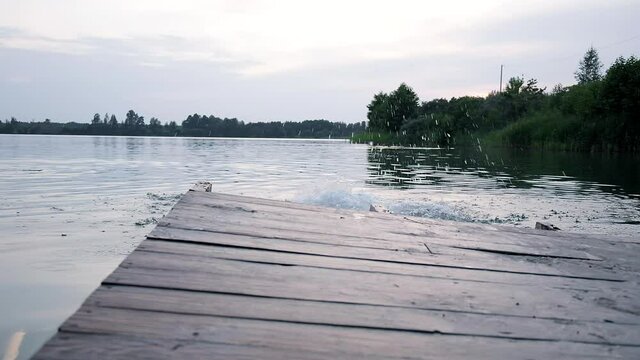 young caucasian male running on wooden dock, jumping in lake with splashes like cannonball. skinny man accelerates and jumps off dilapidated pier in water of pond. summer vacation, rest at nature