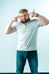 A brutal bearded man holds his hands in the form of pistols near his temples. He is upset, dressed in white polo and jeans on a white background.