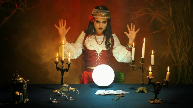 Traditional national pagan fortune telling. Fantasy witch forecaster in old vintage costume predicts fate on fairy glass ball. Backdrop streaming mystical fog, dark gothic room
