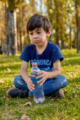 Brunette kid pensively holding a small bottle of water sitting in the middle of the forest at autumn. vertical, blurred background