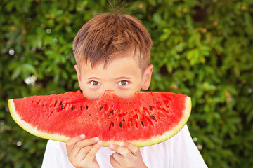 Happy child (boy) is eating a red juicy watermelon. Caucasian kid smiling and having fun. Concept of healthy food, happy childhood, summer vacation. Nature background.