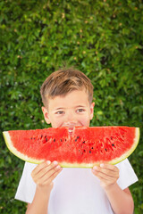 Happy child (boy) is eating a red juicy, tasty  watermelon. Caucasian kid smiling and having fun. Concept of healthy food, happy childhood, summer vacation. Nature background. Copy space.