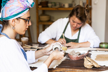 Two young women in aprons in a ceramics studio chatting among themselves and making ceramics, general plan, daylight