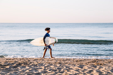 Young afro latino man walking with surfboard along the seashore