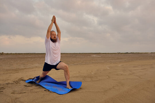Man stands on blue exercise man doing virabhadrasana one pose. Man practices warrior one asana at sea beach during sunrise