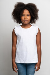 Vertical view of cute little girl with curly hairdo in white T-shirt standing and looking at camera with happy face. Indoor studio shot isolated on white background