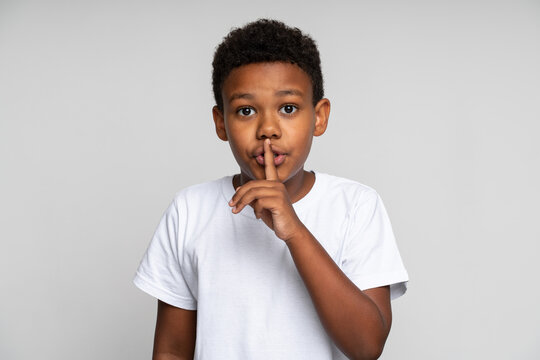 Shh, be quiet. Portrait of funny cute little boy with curly hair in T-shirt making silence gesture with finger on his lips, keeping some secret, child mystery. Studio shot isolated on white background