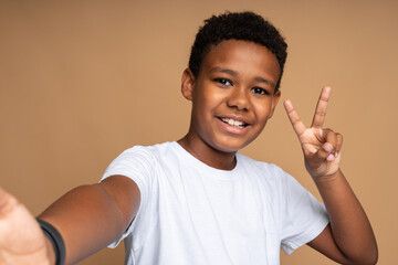 Portrait of happy and victorious little boy in T-shirt showing victory or peace gesture and smiling, doing v sign, number two, cute child enjoying win. Indoor studio shot isolated on beige background © speed300