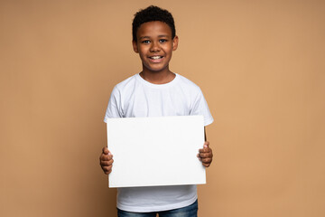 Horizontal view of the cheerful multiracial boy holding a blank white sheet of paper and looking at the camera with toothy smile. Space for text, advertising