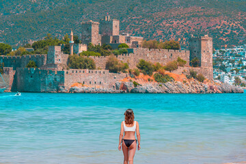 Beautiful girl in black swimsuit walks on a sandy beach -  Panoramic view of Saint Peter Castle (Bodrum castle) and marina - Bodrum, Turkey