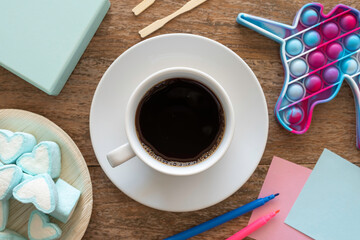 Coffee break concept with cup of coffee, heart shaped marshmallow, notebook paper, felt pens, antistress toy in the form of unicorn over wooden table.