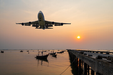 front image commercial passenger aircraft or cargo airplane fly over fishing wood boat floating in the sea at jetty in evening with golden sunset seascape view