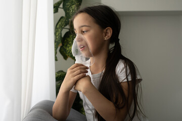 Kid girl makes inhalation with a nebulizer. sick child holding inhalator in hand and breathes through an inhaler at home