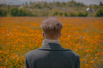 A young guy stands in a field of yellow flowers in a gray coat on a sunny day in summer.