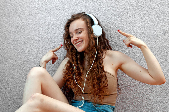 Happy caucasian girl with curly hair and earphones on the floor with a gotele background wall