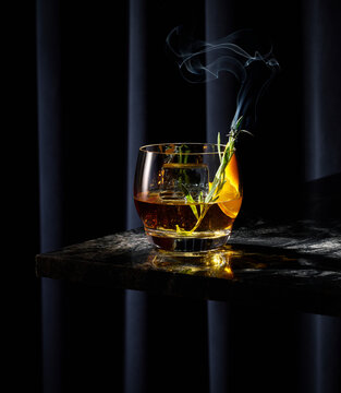 Smoked Whiskey Cocktail with Smoking Lavender Rosemary on Black Granite Bar Table Top with Dark Curtain Background.  Orange peel garnish and large square ice cube.
