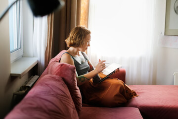 Cute red-haired girl artist with freckles draws on a tablet in a cozy living room real interior,...