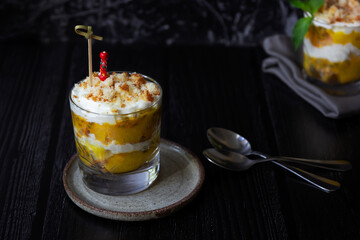 Mango and passion fruit trifle with cream cheese and home made biscuit in a glass on a plate and two small spoons. Dark background, side view