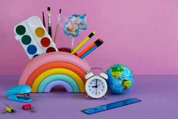 a pencil case in the form of a bright rainbow with a unicorn-shaped lollipop, paints, brushes, pencils, a white alarm clock, a stapler, a ruler and a toy planet on a lilac background - Powered by Adobe