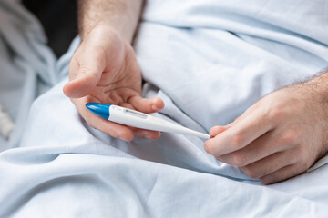 A man lying in bed, holding a thermometer in his hands. Close-up of the hands. The concept of measuring body temperature