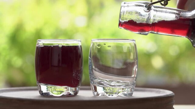 Pouring black elder syrup from a bottle into glasses, with green bokeh in the background