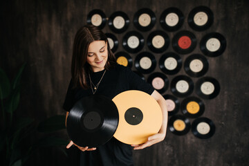 Young woman holding yellow vilyl record in a room with black wall decorated with vintage vinyl...