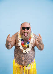 Fototapeta na wymiar Funny bearded man with overweight in decorative grass skirt and flowers garland shows Horns gestures and tongue on light blue background in studio