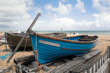 A quaint small blue wooden rowing boat berthed on wooden beams on a pebble beach next to another...