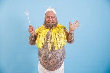 Joyful mature bearded plump man with toy crown, magic stick and yellow cape stands on light blue...
