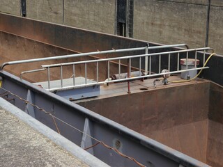 Empty cargo hold of an inland bulk carrier, the walls are rusted, a walkway connects the side walls, railings and various hoses and lines are installed.  The ship is in the low position of a lock