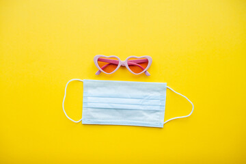 Pink heart shaped sunglasses and protective medical mask on bright yellow background. High quality photo