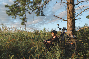 Man in helmet sitting on grassy ground near bicycle and coniferous tree and resting during ride in countryside