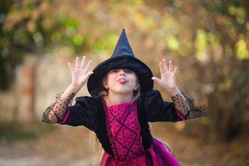 A portrait of a little girl in a witch costume hiding behind a black cap and showing her tongue. Halloween holiday. Copy space