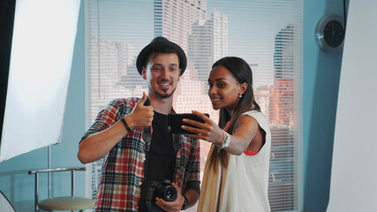 Beautiful african model making selfie with handsome photographer in professional studio photo shoot. There is soft box in the background.