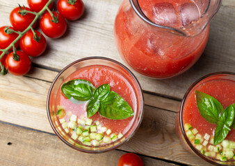 Top view of gazpacho soup in glass glasses and a jug of ice on a wooden table. Cherry tomato branch, basil leaves and fresh cucumber, sliced brunois