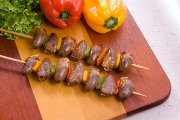 Tábua de madeira com espetos de churrasco com coração de galinha, pimentões e alface em mesa branca.